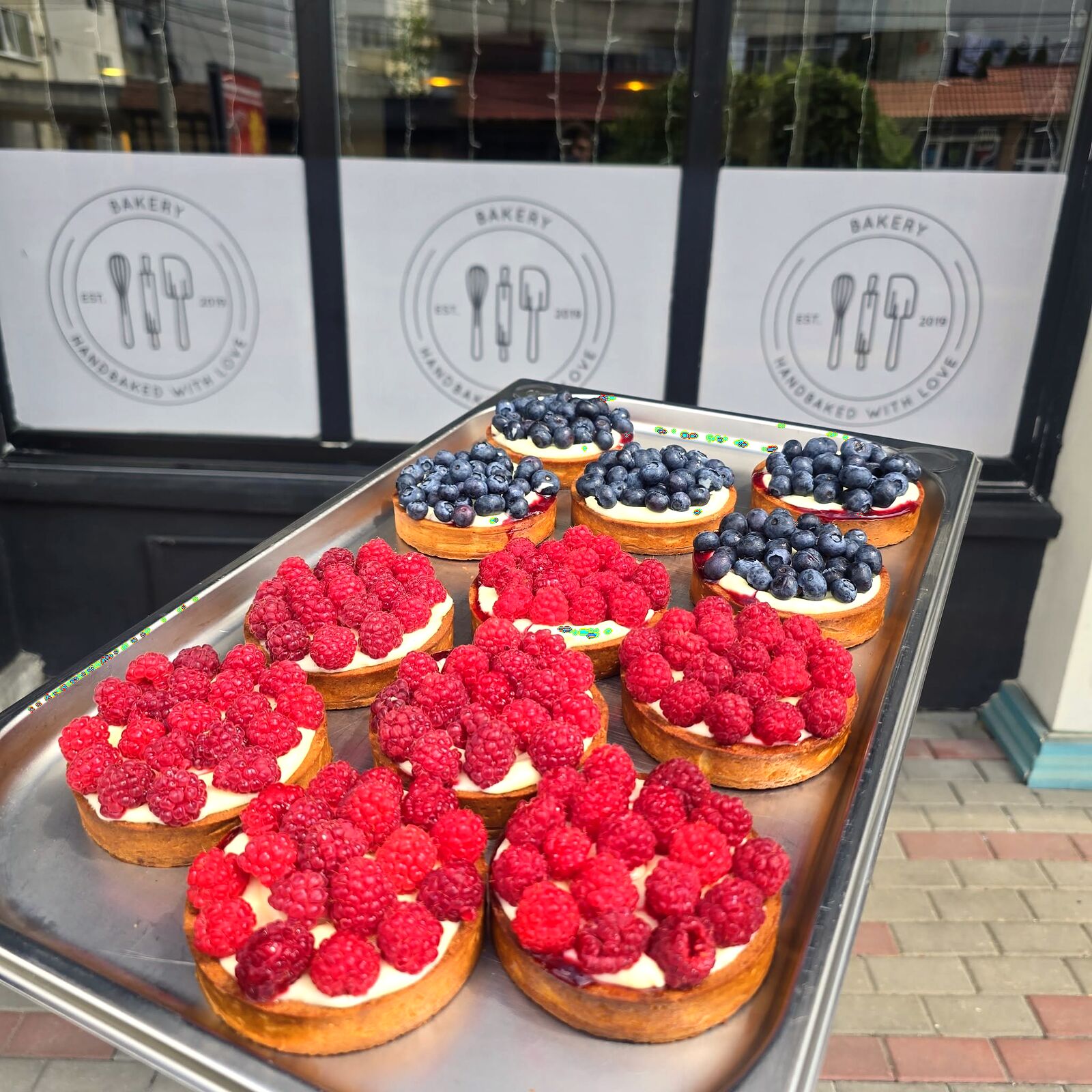 Tarte cu cremă de vanilie, zmeură și afine, pe tavă, în fața geamurilor cu sigla Bakery